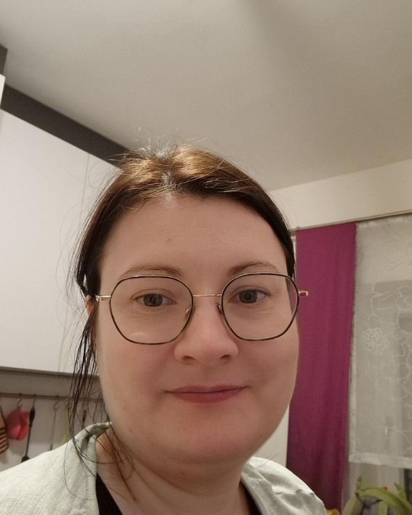 Das Bild zeigt Martina, die in einer Küche mit weißer Arbeitsfläche und lila Vorhängen im Hintergrund steht.