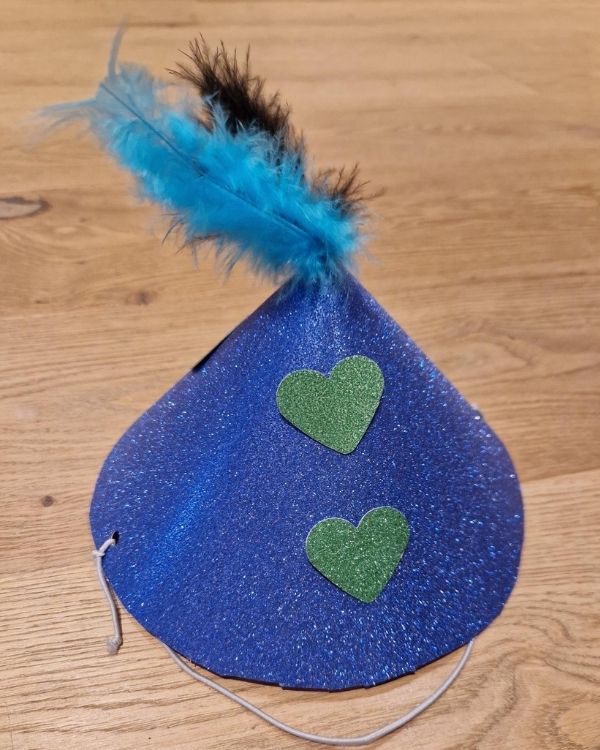 Selbstgebastelte Partyhut-Mütze in blauem Glitzerstoff mit zwei grünen Glitzerherzen und einer blauen Feder mit schwarzem Akzent an der Spitze.